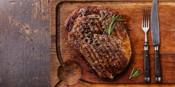 Grilled Black Angus Steak Ribeye and fork and knife on meat cutting board on dark wooden background